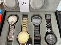 Four wristwatches displayed in a grey velvet watch holder case. Watches include a silver-tone with crystal details, a gold-tone heavy rhinestone design, a black-tone crystal accented watch, and a simple black stainless watch.