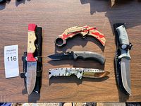 Five knives on wooden table, including wooden handle fixed blade, karambit folding knife, black fixed blade, silver folding knife with holes, large fixed blade.