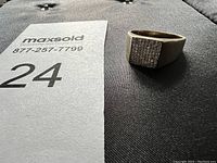 Ring displayed on a dark fabric surface beside a MaxSold number card, showing the front rectangular pavé stone setting.