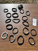 Overview photo of all 19 bracelets spread on wooden table surface. Shows a variety of black beaded bracelets, braided leather bracelets, and a translucent white bead bracelet.