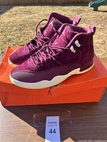 Pair of burgundy Air Jordan 12 Retro sneakers on top of orange shoebox, displayed outdoors showing general appearance and style.