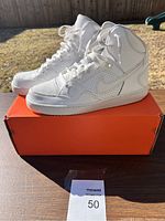 Side angle showing the white Nike high-top sneaker pair on orange Nike shoebox with natural outdoor lighting.