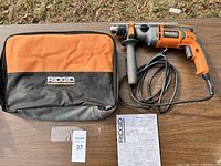 Photo showing the Ridgid hammer drill, branded orange and black soft carry bag, and operator's manual laid on a flat surface outdoors.