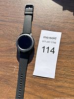 Samsung Galaxy Watch front view showing black round watch face and black silicone band placed on wooden surface with auction lot paper beside it.