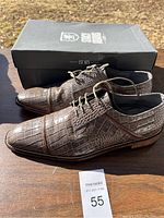 Two brown faux crocodile leather patterned dress shoes in lace-up style with wooden-look sole and shoe box in background.