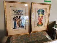 Two framed and matted paintings with Ancient Egyptian themes on papyrus. One features three women with musical instruments; the other is a head portrait of an Egyptian figure. Gold-tone frames and cream mattes shown.