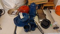 Photo showing blue enamelled Dutch oven, plates, coffee cups and pot, mandolin slicer, cake pan, and thermometer alongside grilling spatulas and a black plastic basket.