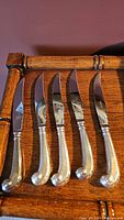 Set of five silver-handled cutting knives with curved scroll handles and shiny blades placed on wooden surface.