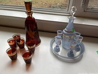 Photo showing red glass decanter with six matching red glasses featuring gold trim and etched bird and bull rush designs, alongside frosted decanter set with five glasses and tray.