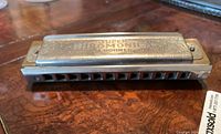 Full view of a metallic chromatic harmonica with engraved 'SUPER CHROMONICA HOHNER' branding, showing the numbered holes and overall structure.