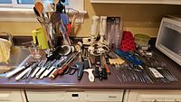 Overview of kitchen counter with various cutlery, knives, forks, spoons, spatulas, handheld blenders, and kitchen gadgets lined up.