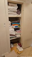 Full linen cupboard showing neatly stacked white and colored towels, bed sheets, and fabric items on four shelves and floor.