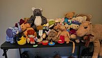 Photo of the complete collection of plush toys including teddy bears, a camel, elephant, and other assorted animals on a black table.
