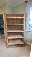 Front view of mid century style rattan wicker shelving unit placed near window in room, showing five shelves and woven design on back and sides.