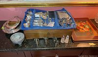Full view of cutlery set in blue fabric-lined wood storage box placed on counter with other silver plated serving pieces, tray, and napkin rings.
