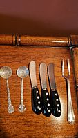 Photo showing two Mexican souvenir spoons marked 925 along with three black-handled butter cutters and one silver-toned cocktail fork laid out on wood surface.