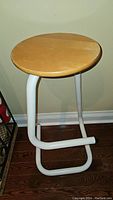 Photo of a round wooden top stool with white tubular steel paperclip-style base against a plain wall, showing seat and base design.