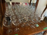 Photo showing a collection of multiple clear glass wine glasses arranged on a wooden cabinet surface with Trudeau style glasses in front and Libby style glasses behind.