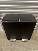 Front view of the closed dual compartment black step trash can showing two separate foot pedals and lids.