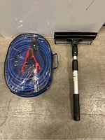 Plastic zipper pouch containing coiled blue jumper cables with red and black clamps and black handled window squeegee placed side by side on the floor.