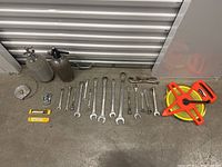 Overview of various hand tools laid out on a concrete floor in front of a metal shutter. Includes measuring tapes, wrenches, siphon bottles, chain pipe wrench, and batteries.