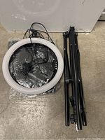 Photo shows the ring light and the folded tripod stand placed side by side on the ground. The ring light has an attached power cable with a two-prong plug and control buttons visible.