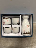 Top-down view of porcelain sake carafe and four cups arranged in blue box with white insert.