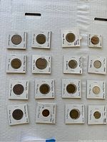 Top view of various foreign coins individually placed in white protective holders with handwritten details about origin, year and denomination in a grid format