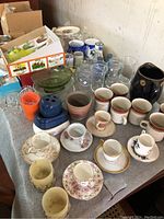 Wide view of the eclectic glassware collection on a table, five small plates (one broken), various teacups, saucers, shot glasses, mugs, ramekins, and other glassware.