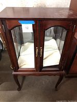 Front view of the Powell glass display cabinet showing wood frame, glass panel doors with brass handles, and fabric covering inside base.
