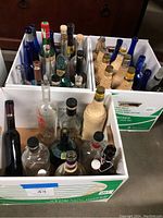 Multiple empty liquor bottles arranged in cardboard boxes, showing range of colors including cobalt blue, clear, and woven coverings.