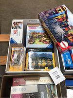 Multiple jigsaw puzzle boxes stacked in an open box including Ravensburger 1000 piece and 500 piece puzzles, Antex 500 piece puzzle, some loose puzzle pieces in plastic bag.