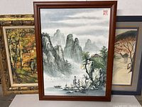 Framed Asian-style landscape painting showing misty mountains, pine trees, boats on water, and rocky terrain with red artist stamp