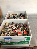 Two large white storage boxes filled with numerous matchbooks of various brands and locations, placed on a gray surface.