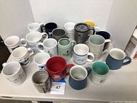 Top and front view of 21 assorted souvenir coffee mugs in varied colors and printed designs, arranged closely together.