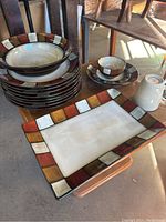 Set of stacked plates, bowls, mugs, and a rectangular serving platter all with colorful bordered pattern on white ceramic.