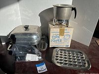 Lot contents showing vintage metal popcorn popper on its original box, electric skillet with lid behind, and an electric wine opener in a plastic bag.