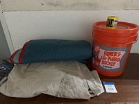 Lot comprising a blue moving blanket, large white canvas drop cloth with dirt, an orange 5-gallon bucket from Home Depot, and a small yellow tape roll.