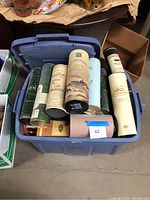 Blue plastic bin filled with tall, cylindrical whiskey canisters in various colors and brands, some paper/cardboard and some metal, approx. 9-15 inches tall, packed upright.
