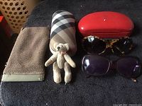 Two pairs of vintage sunglasses, one with tortoiseshell frame and one with dark rectangular frame, both show wear; one arm on dark pair broken. Two vintage sunglass cases, one hard red and one beige plaid soft case. Small vintage stuffed bear toy included.