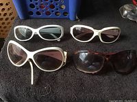 Four pairs of vintage women's sunglasses laid out on black fabric background showing white and tortoiseshell large rounded frames with tinted lenses.