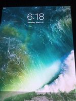 iPad Mini powered on showing lock screen with time and date, typical Apple iPad wallpaper with green and blue waves.