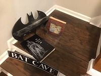 Photo of black metal Batman logo shelf, Bat Cave sign, and two Batman-themed books on wooden floor