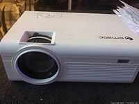 View of the front and top side of the white Gematic projector, showing lens and product branding