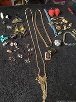 Overview of the lot laid out on a black surface showing a collection of jewelry items including necklaces, earrings, a watch, rings, bracelets, and pins.