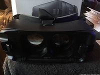 Photo showing the front view of the Samsung Gear VR headset with visible dual lenses and strap, placed on a fabric surface.