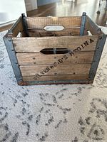 Front angle view showing the wooden slats, metal reinforced corners, and cut-out handles with stencil text on one side.