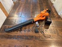 Black and Decker leaf blower with charger and power adapter on wooden table, showing full assembly with blower tube attached.