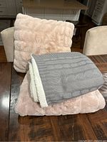 Photo showing plush light pink throw pillow propped up on table with two folded blankets, a pink plush blanket beneath and a grey and white knitted blanket on top.