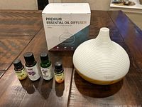 Essential oil diffuser with wood-tone base and white ribbed ceramic top, along with four bottles of essential oils in front of the box.